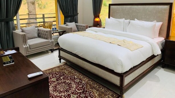 A spacious bedroom with a large bed, a sitting area with a chair and a small table, and a view of the outdoors through large windows.