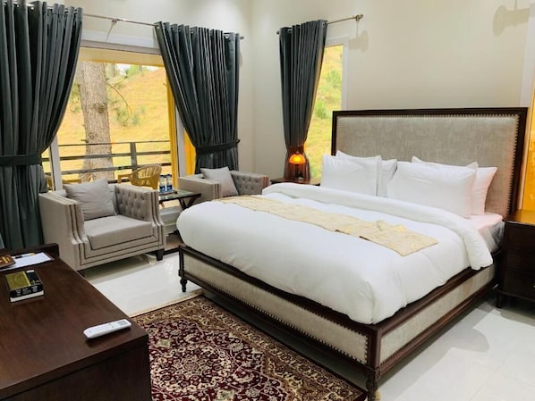 A spacious bedroom with a large bed, a sitting area with a chair and a small table, and a view of the outdoors through large windows.