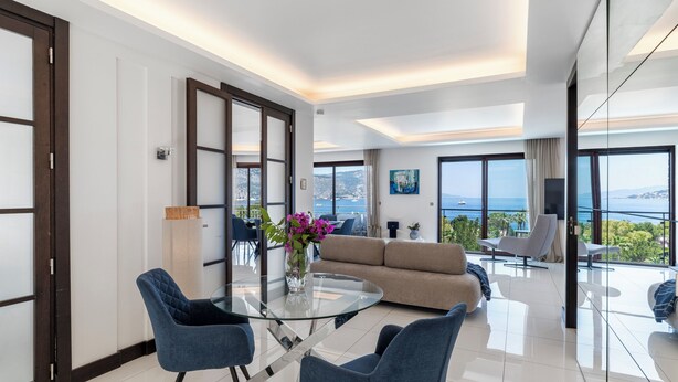 Une chambre d’hôtel moderne avec une table à manger en verre, des chaises bleues, un canapé beige et une vue sur la mer.