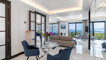 Een moderne hotelkamer met een glazen eettafel, blauwe stoelen, een beige bank en uitzicht op zee.