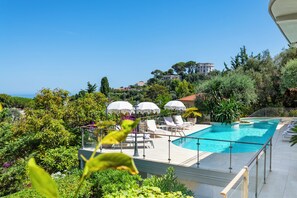 A swimming pool with lounge chairs and umbrellas, surrounded by greenery and a view of a hill with buildings. - Exclusive Riviera Villa for 16 Guests | Pool & Ocean View (Saint-Jean-Cap-Ferrat)