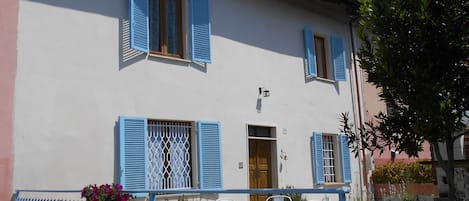 A two-story building with blue shutters, a balcony with a metal railing, and potted plants.