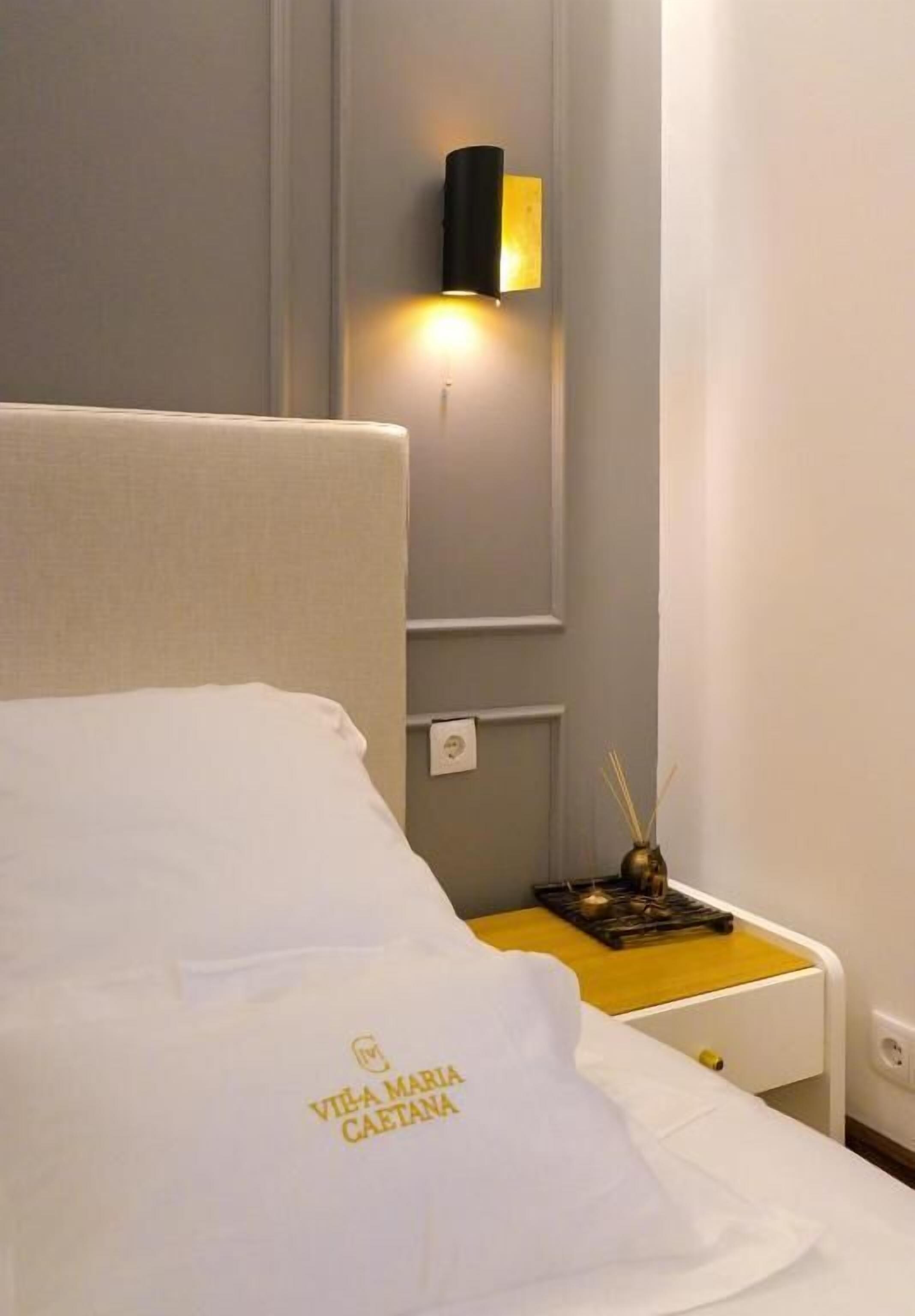 A hotel room with a bed, a nightstand, a wall-mounted light, and a small table with a candle.