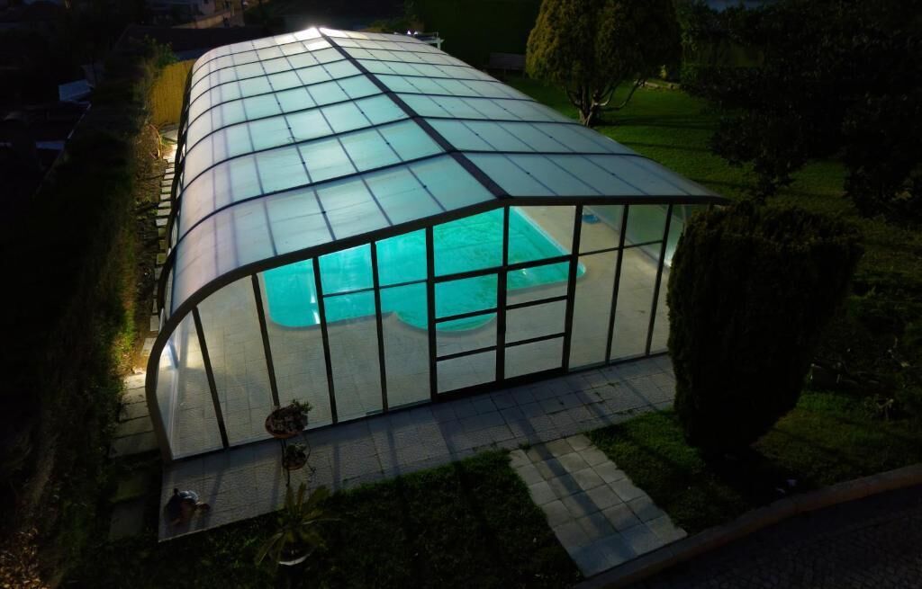 A glass-enclosed swimming pool with a curved roof, surrounded by a paved deck and greenery.