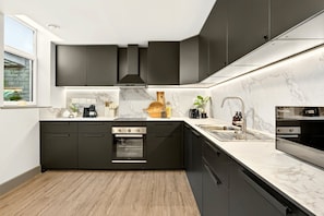 A modern kitchen with dark cabinetry, a built-in oven, a sink, and a marble backsplash. - Guestready - The Bridge - Wealdstone (Harrow)