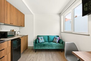 A compact kitchen with wooden cabinets, a microwave, and a dishwasher. A teal sofa with floral cushions is positioned against the wall, and a window provides a view of the outside. - Guestready - The Bridge - Wealdstone (Harrow)