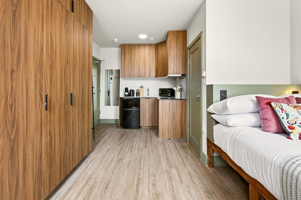 A compact room with a kitchenette, a bed with pillows, and a wooden wardrobe.