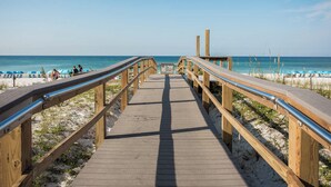 Ein hölzerner Steg führt zu einem Strand mit blauen Sonnenschirmen und Liegestühlen.