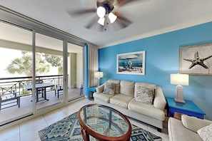 A room with a sliding glass door leading to an outdoor area, a ceiling fan, a sofa with cushions, a glass coffee table, and a painting on the wall. - Azure #208 | Near Beach, Beautiful Views + Amenities (Fort Walton Beach)