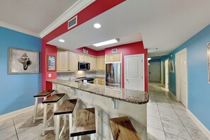 A kitchen with a central island, bar stools, wooden cabinets, a microwave, and a refrigerator. - Azure #208 | Near Beach, Beautiful Views + Amenities (Fort Walton Beach)