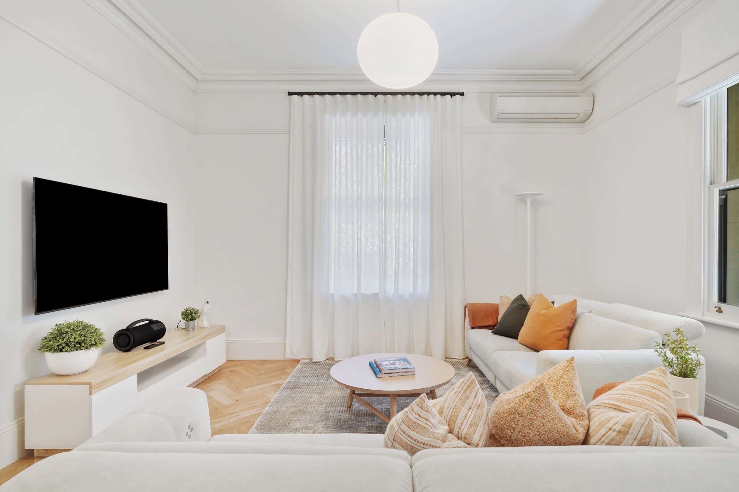 A modern living room with a flat-screen TV, a white sofa, a coffee table, and a large window with sheer curtains.