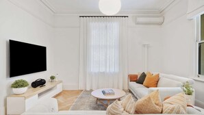 Uma sala de estar moderna com televisão de tela plana, um sofá branco, uma mesa de centro e uma janela grande com cortinas leves.