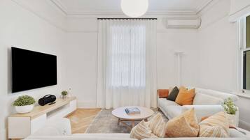 A modern living room with a flat-screen TV, a white sofa, a coffee table, and a large window with sheer curtains.