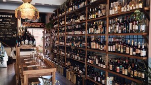 Um bar com prateleiras de madeira repletas de garrafas diversas, um menu em lousa e um candeeiro suspenso.
