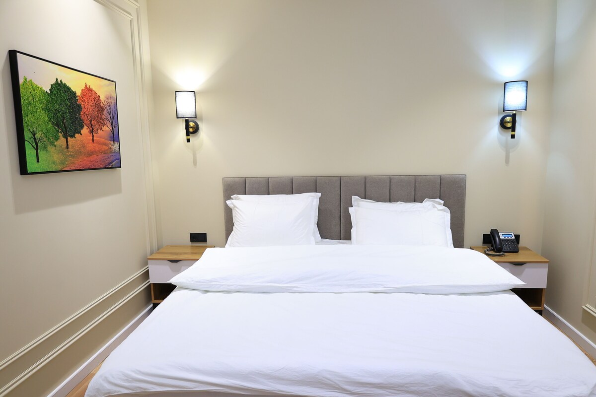 A hotel room with a large bed, two bedside tables, a painting on the wall, and two wall-mounted lights.