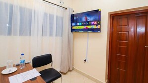 Ett hotellrum med en tv som är monterad på väggen och visar en sportkanal, en dörr av trä och ett fönster med tunna, genomskinliga gardiner.