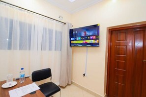 Habitación de hotel con un televisor montado en la pared mostrando un canal deportivo, una puerta de madera y una ventana con cortinas de gasa.