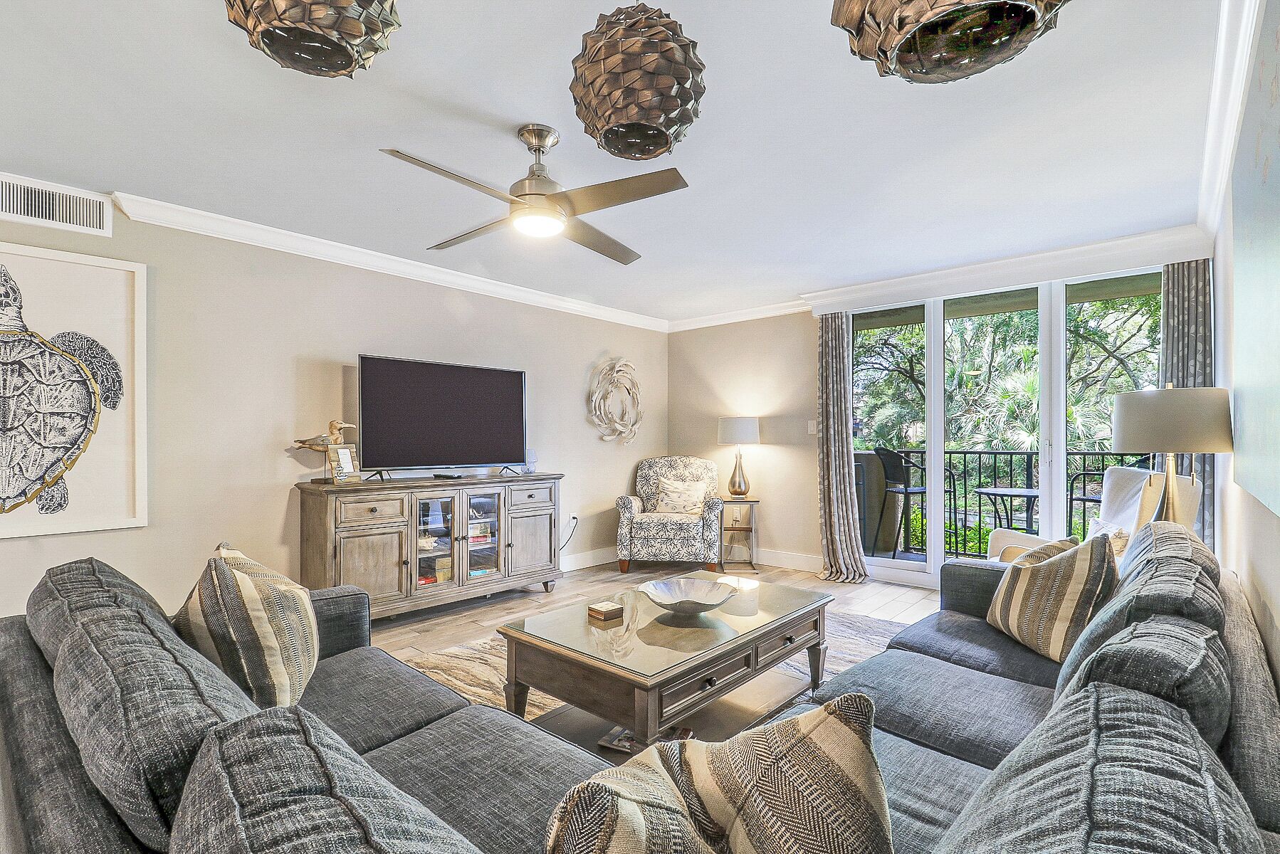 A living room with a sofa set, a television, a ceiling fan, and a window with a view of trees.