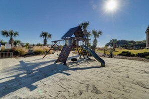 Ein hölzerner Spielplatz mit Rutsche und Kletterwand, umgeben von Sand und Palmen.