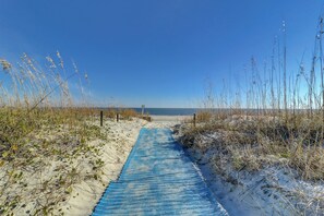 Ein Strandweg, der zum Meer führt, umgeben von Sand und vereinzelter Vegetation.