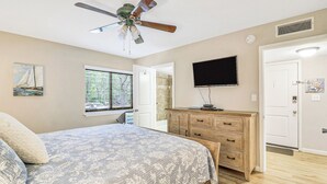 Een slaapkamer met een bed, een plafondventilator, een televisie, een dressoir en een raam met jaloezieën.
