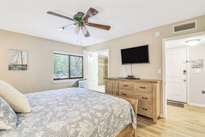 Ein Schlafzimmer mit einem Bett, einem Deckenventilator, einem Fernseher, einem Schminktisch und einem Fenster mit Jalousien.