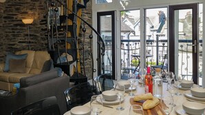 A dining area with a spiral staircase, a sofa, dining tables set with plates and glasses, and a view of the street outside. - Hillside Cottage (Windermere)