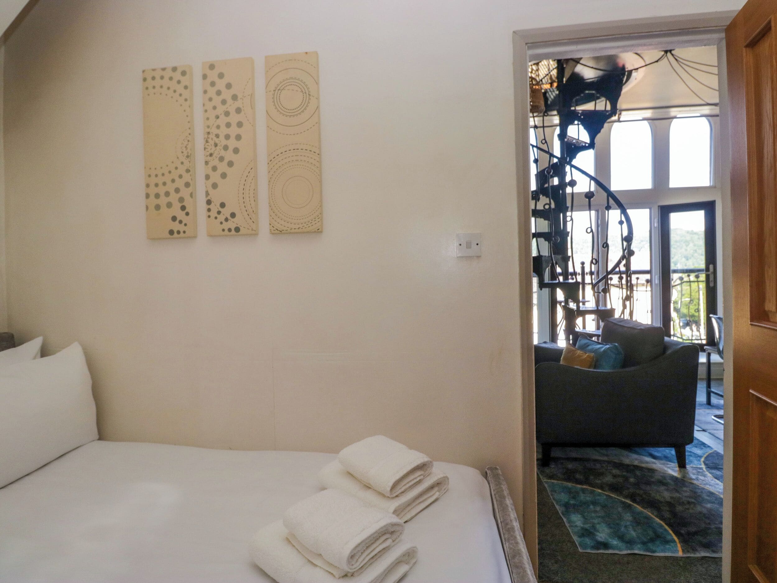 A bedroom with a bed, two wall art pieces, a view of a balcony with a chair, and a staircase.
