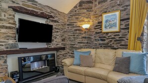 A living room with a stone wall, a flat-screen TV mounted above a fireplace, a sofa with cushions, and a lamp. - Hillside Cottage (Windermere)