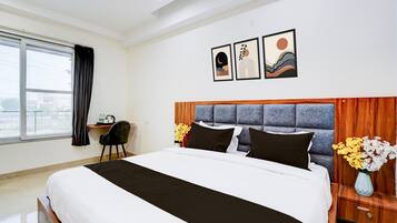 A hotel room with a large bed, a desk, a chair, a window, and artwork on the wall.