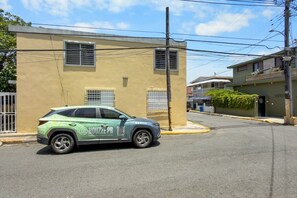 A light green SUV parked on a street in front of a beige building with a window and a door. - Casa Costa Brava | 8 Room - 16 Guest @ San Juan (San Juan de Puerto Rico)