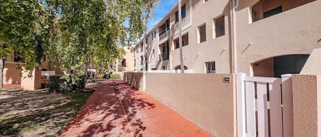 Un sendero que lleva a un edificio de fachada color crema, con una entrada protegida por una reja y un cielo despejado.