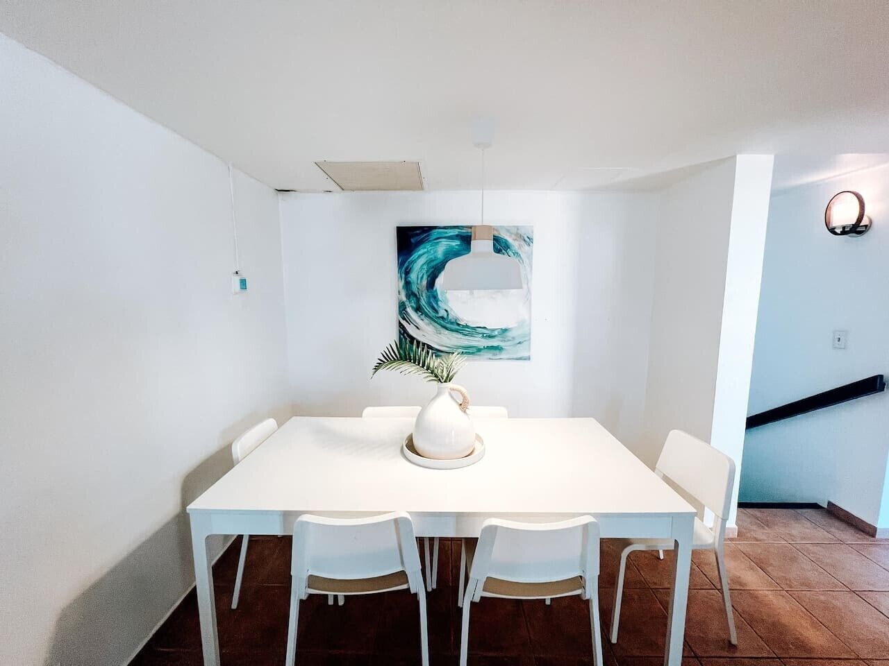 A dining area with a white table and chairs, a painting on the wall, and a staircase in the background.