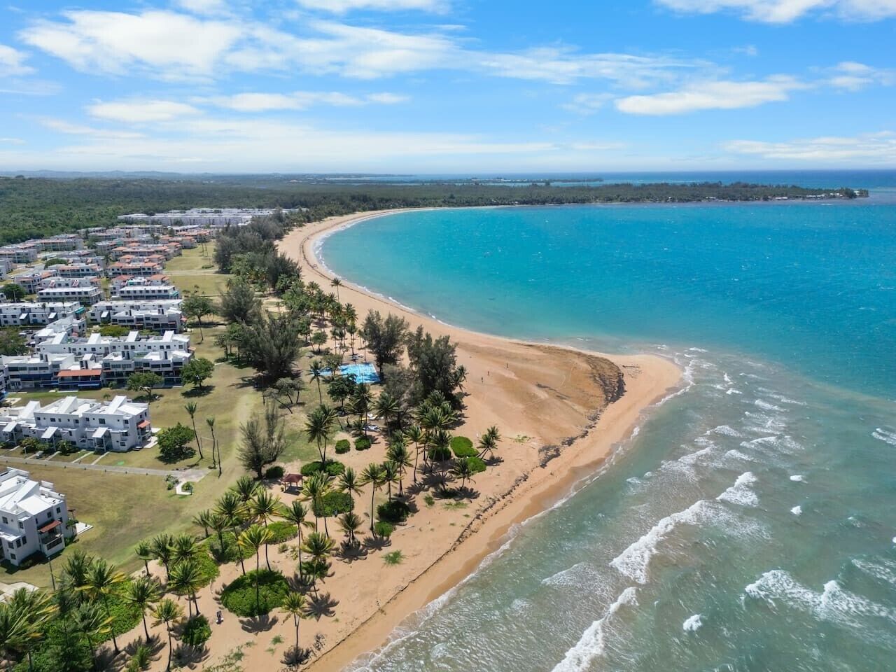 An aerial view of a coastal area with a beach, a residential complex, and a clear sky.