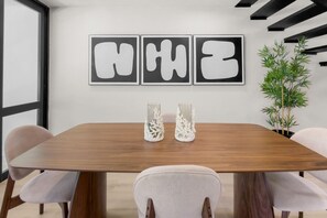 A conference room with a wooden table, white chairs, three abstract paintings, and two glass vases with white coral-like structures. - Villa Marena | Playas del Yunque Modern Apt (Rio Grande)