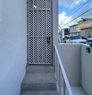 A narrow stairway leading to a gated entrance with a metal lattice door, adjacent to a building with a white exterior and a visible street with parked cars. - Central San Juan 3BR Apt • Sleeps 8 (San Juan de Puerto Rico)