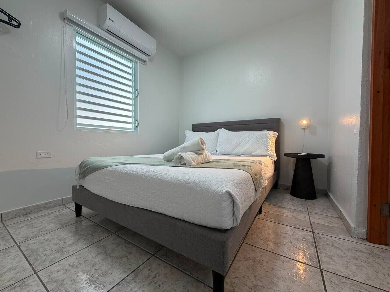 Une chambre à coucher comprenant un lit, une table de chevet, une fenêtre avec des stores et un appareil de climatisation.