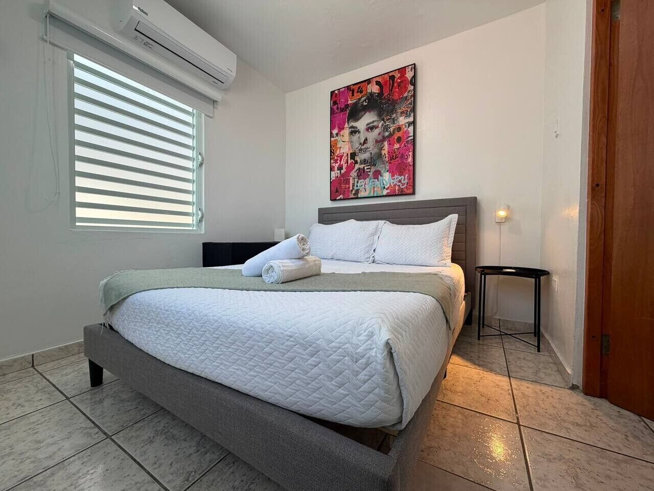 Ein Schlafzimmer mit einem Bett, einem Nachttisch, einer Wand-Klimaanlage und einem Fenster mit Jalousien.