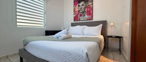 A bedroom with a bed, a bedside table, a wall-mounted air conditioner, and a window with blinds.