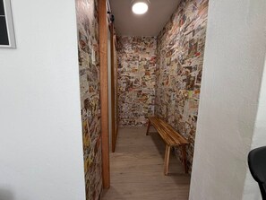 Ein schmaler Flur mit einer hölzernen Bank, Wänden, die mit Zeitungsartikeln tapeziert sind, und einer Deckenleuchte.
