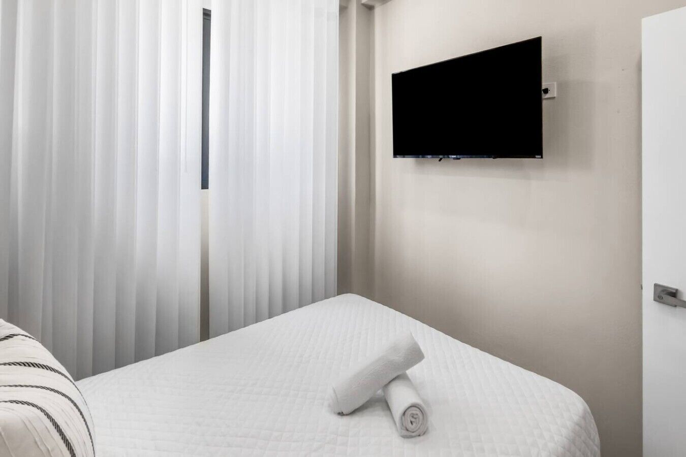 Una habitación de hotel con una cama, un televisor montado en la pared y un ventanal con persianas verticales.