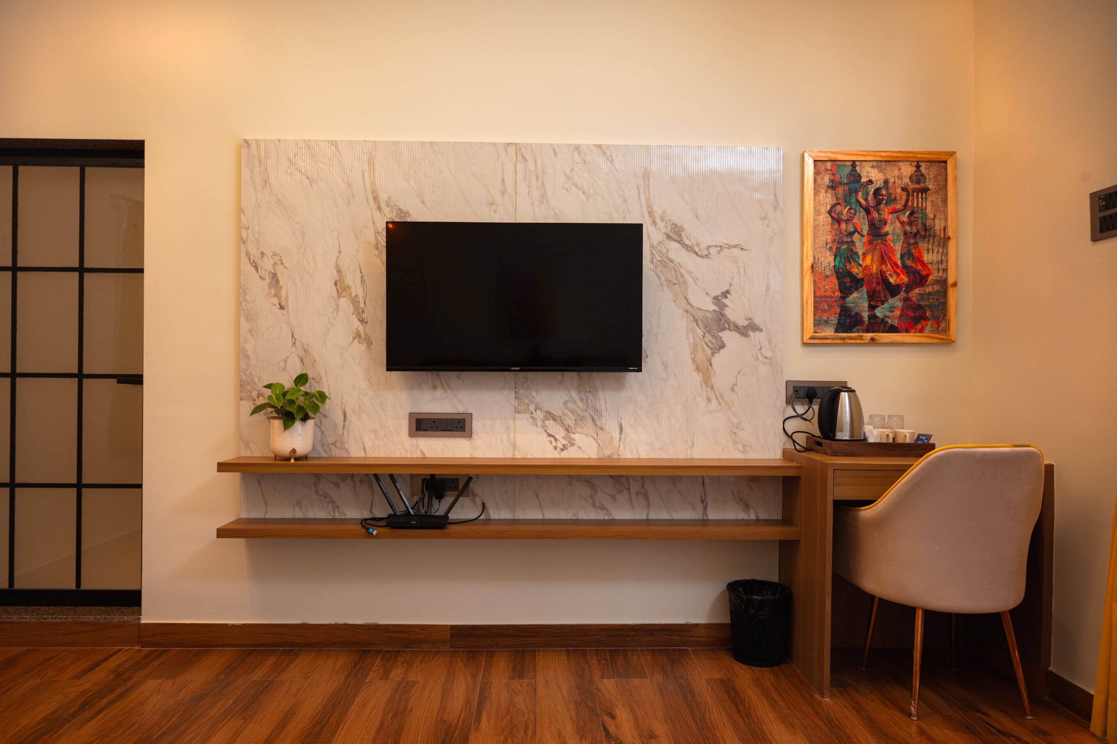 A modern room with a flat-screen TV mounted on a marble wall, a wooden desk with a chair, a potted plant, and a painting.