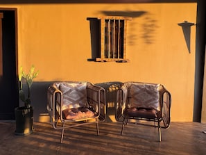 Two wicker chairs with cushions, a potted plant, and a window with a wooden frame against a yellow wall.