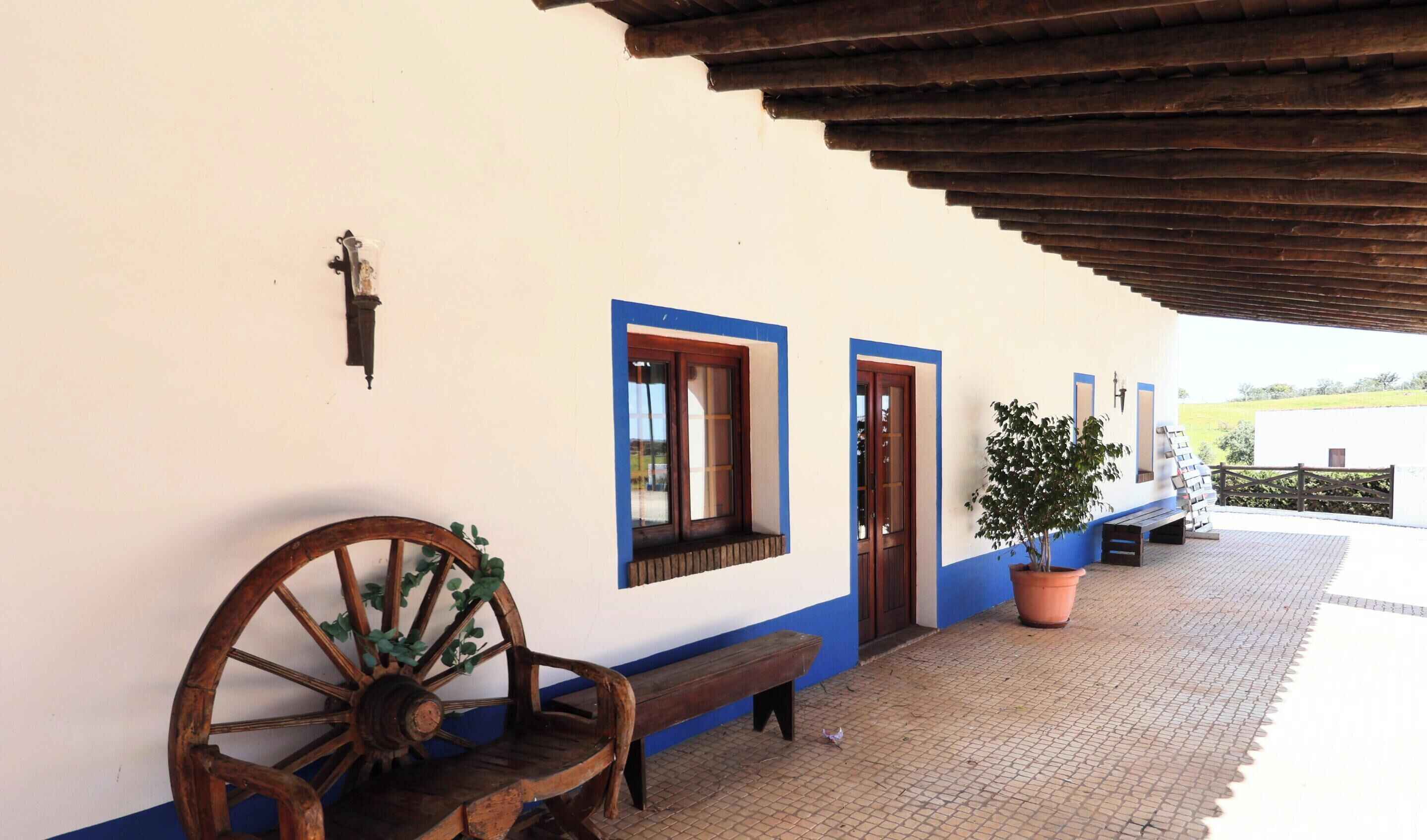 A covered outdoor area with a bench, a wooden wheel, a potted plant, and a wall with a door and windows.