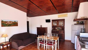 Una sala de estar con sofá, una mesa y sillas, televisor, mueble con puertas de cristal y una zona de cocina con nevera y lavaplatos.