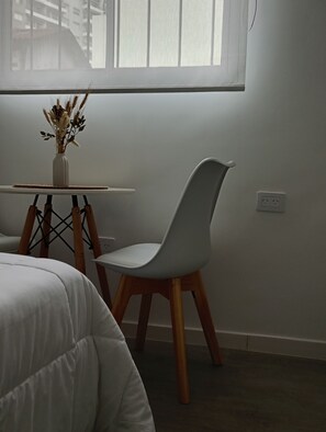 A room with a bed, a chair, a small table with a vase, and a window with blinds. - Acogedor departamento en Villa Urquiza (Buenos Aires)