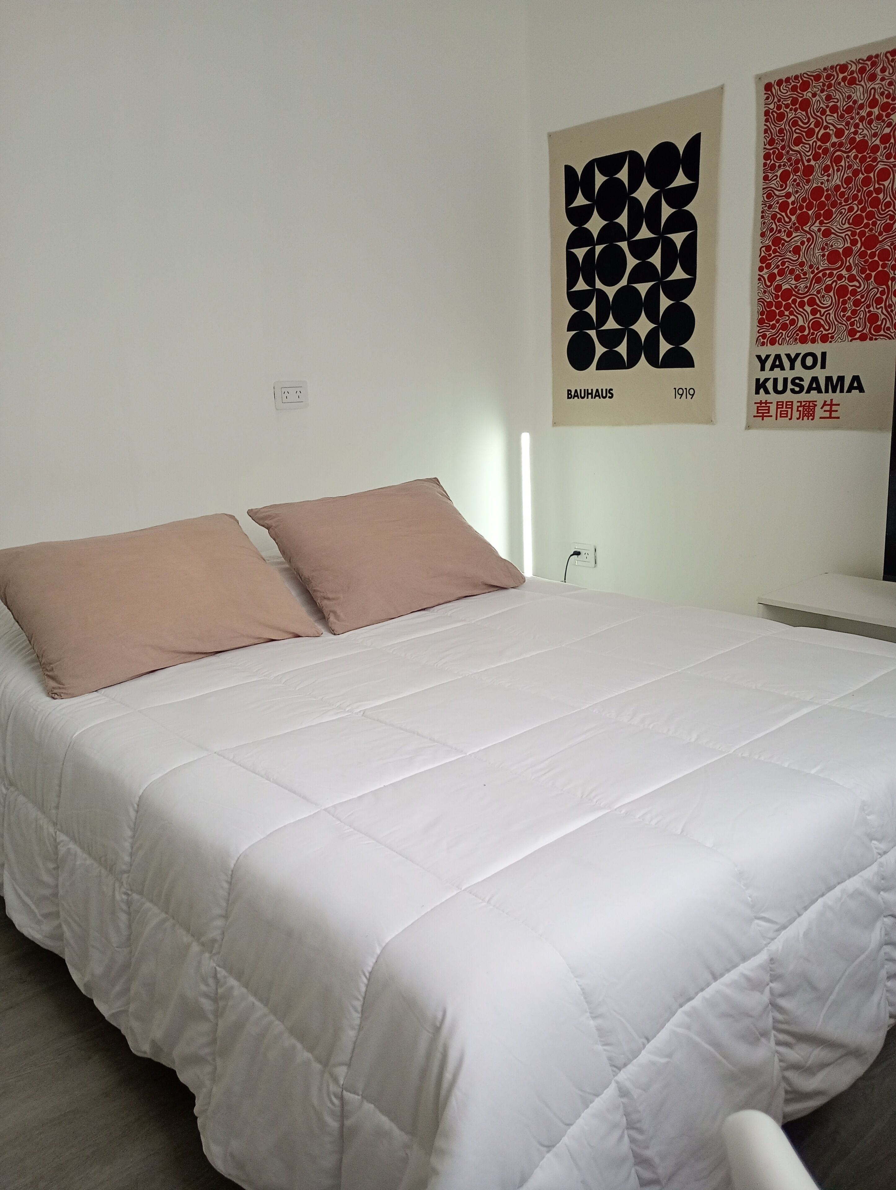 A neatly made bed with a white duvet and two pillows, a wall socket, and two posters.