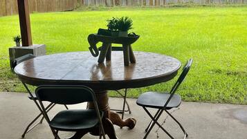 A patio with a round table and chairs, a small potted plant, and a view of a grassy yard and a wooden fence.