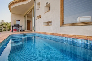 Ein Swimmingpool mit einer Fliesenleiste, eine Terrasse mit Tisch und Stühlen und ein Gebäude mit großem Fenster und Eingangstür.