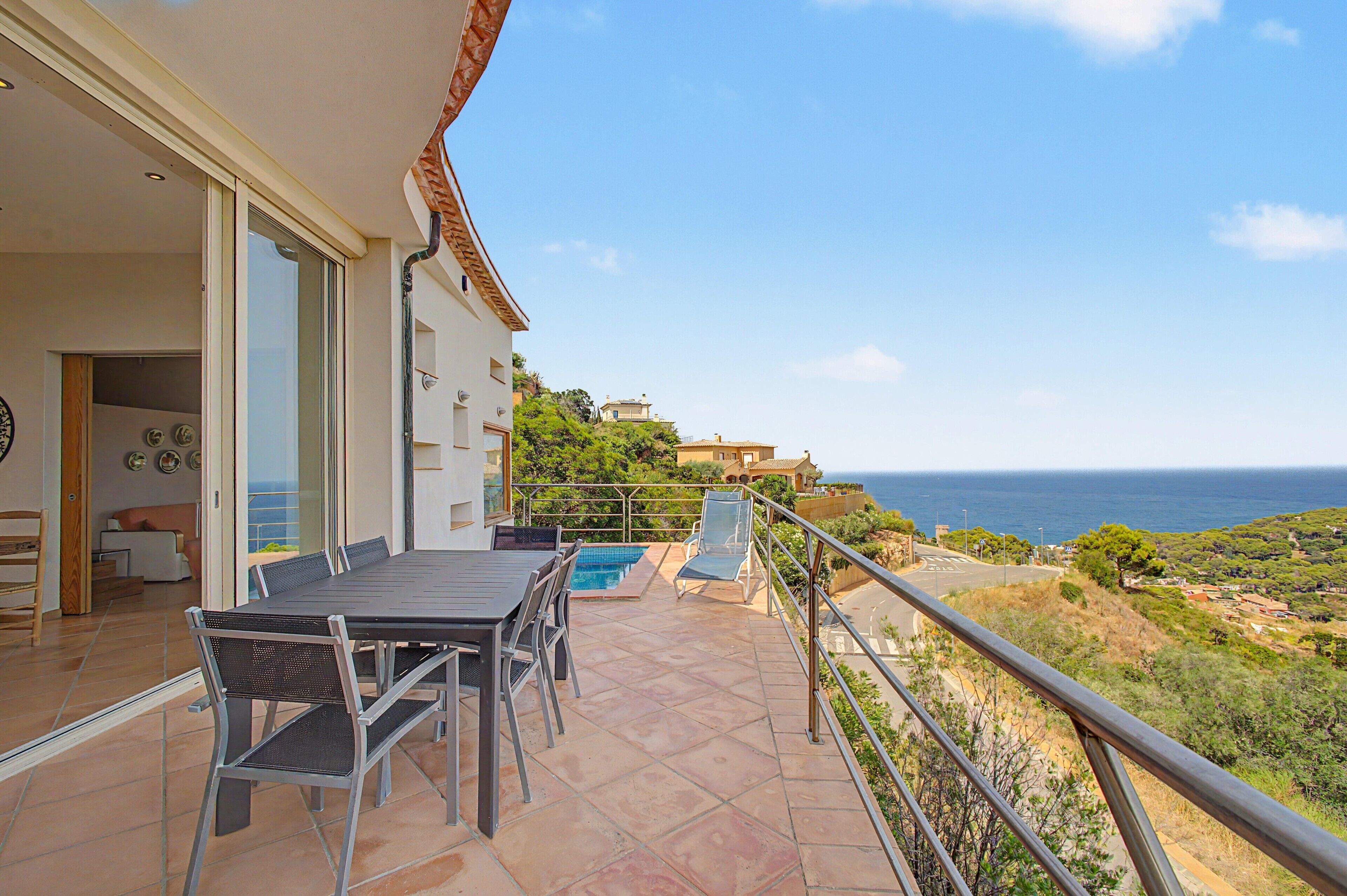 A balcony with a dining table and chairs, a pool, and a view of the sea.